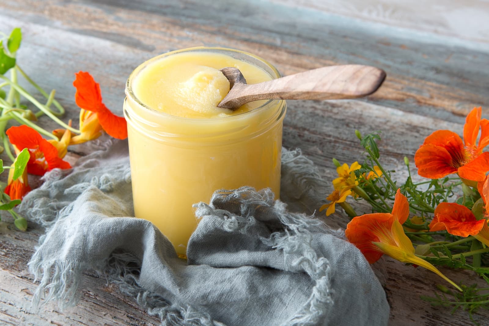 Glass jar of melted ghee with a wooden spoon, placed on rustic wood with flowers and cloth, illustrating an ancient wisdom on ghee article feature image.